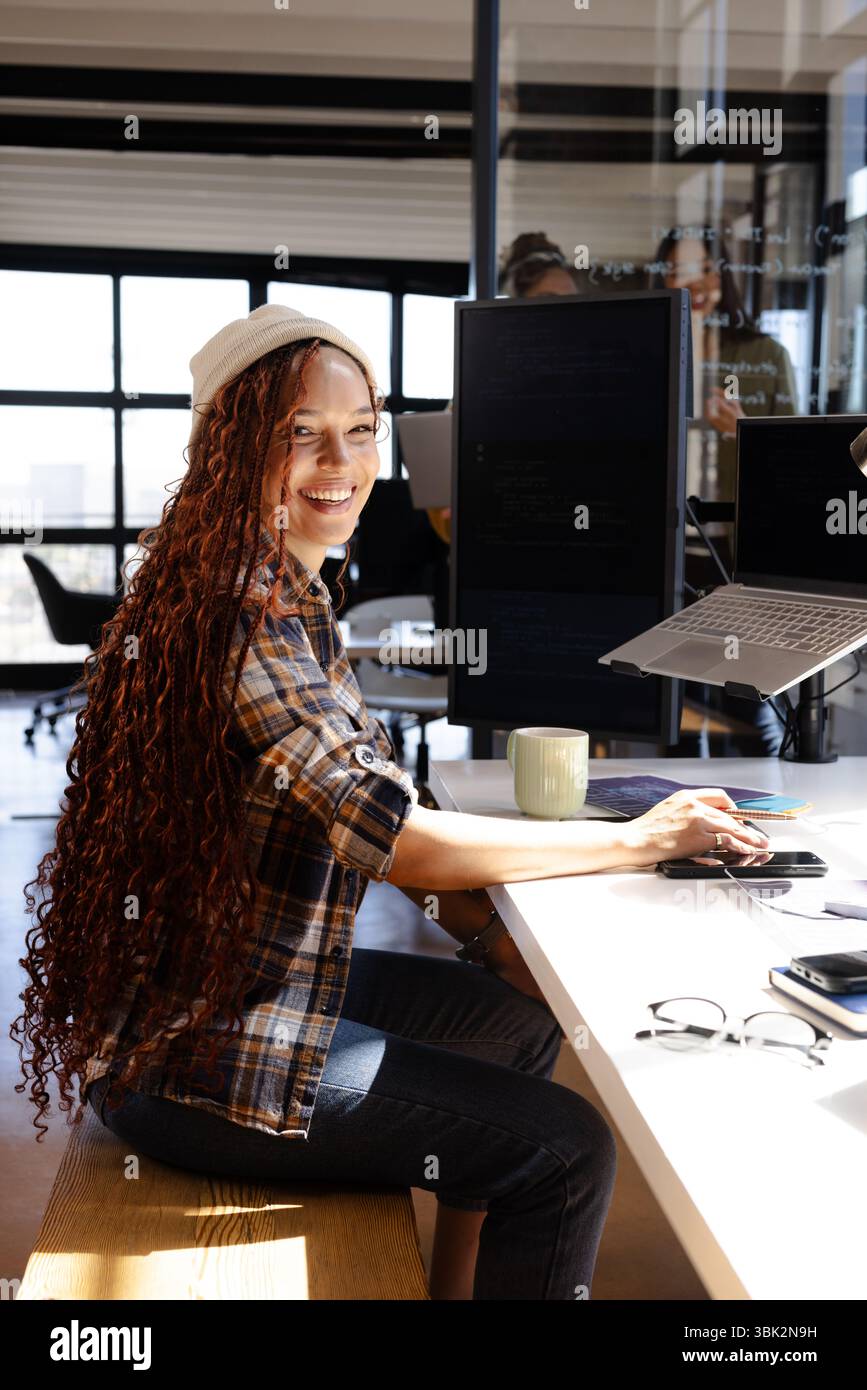 Femme souriante codant au bureau avec ordinateur portable et deux moniteurs dans le bureau Banque D'Images