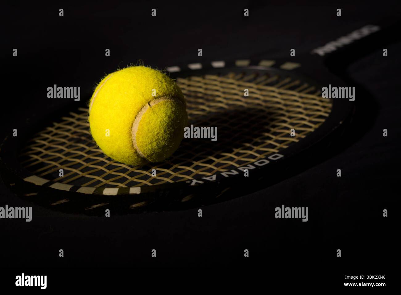 Gros plan d'une balle de tennis jaune éclatante reposant sur les cordes d'une raquette de tennis classique, sur un fond sombre et atmosphérique. Banque D'Images