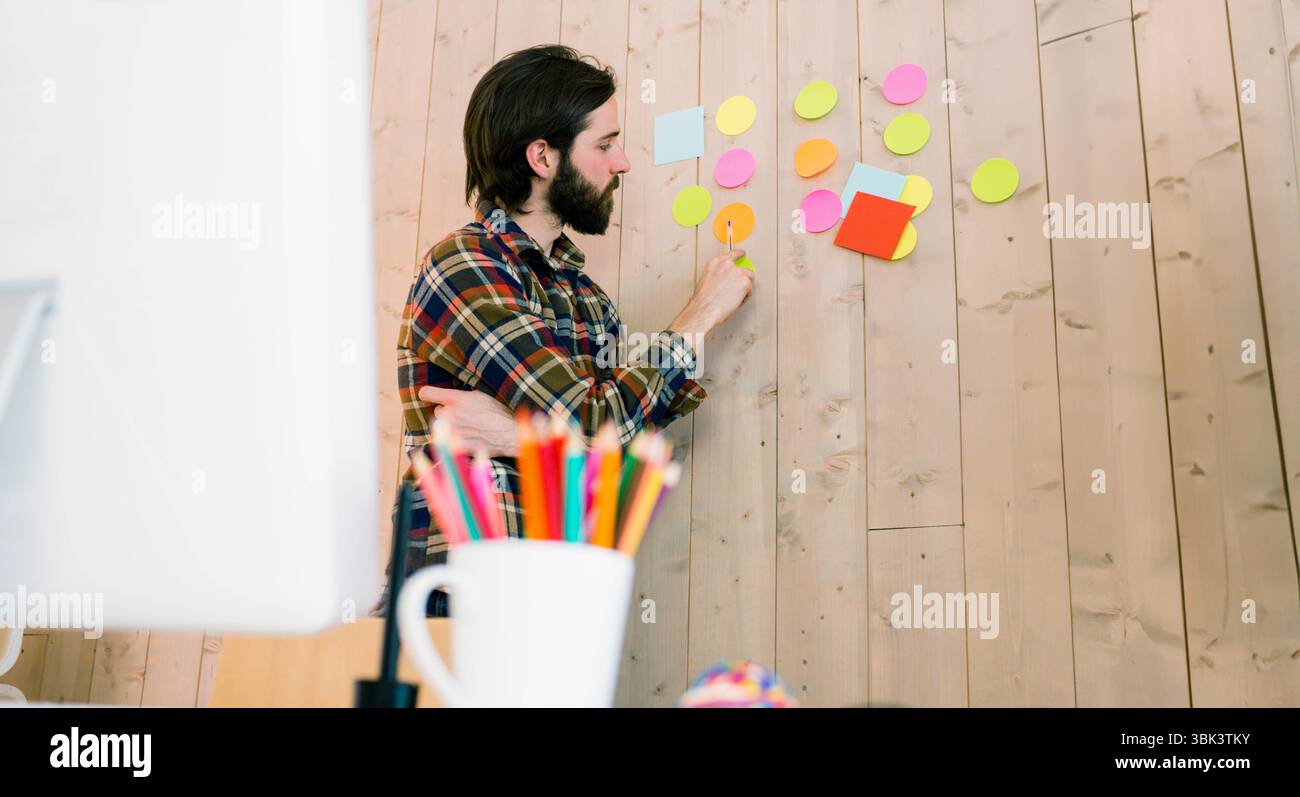 Homme plaçant des notes adhésives rondes et carrées colorées sur le mur d'accent en bois dans un espace de travail de bureau moderne Banque D'Images