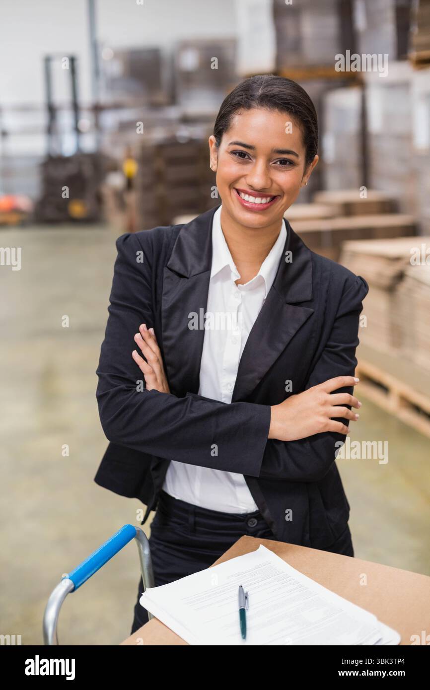 Femme portant un blazer debout avec les bras croisés dans l'entrepôt à côté du chariot à roulettes tenant la paperasse Banque D'Images