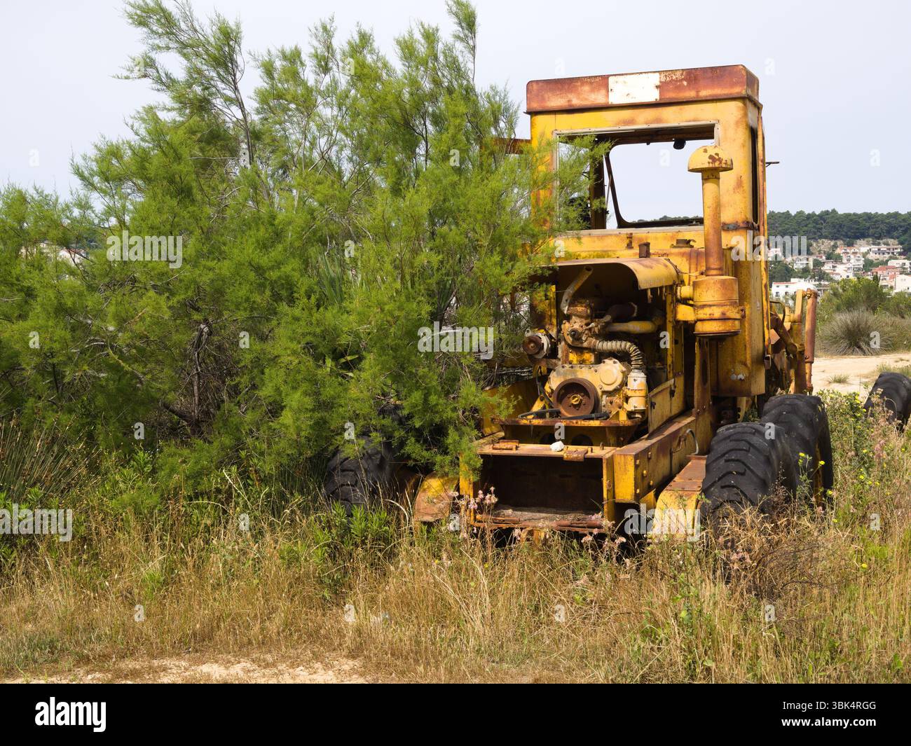 Tracteur jaune altéré avec des restes de moteur visibles abandonnés dans un champ de campagne sec à côté d'un buisson vert vibrant. Banque D'Images