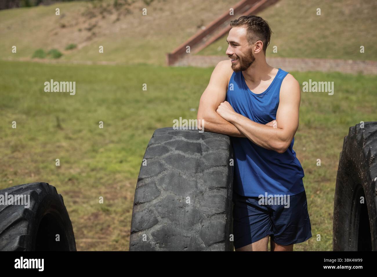 Fit homme dans les vêtements de sport penchant avec les bras croisés sur le pneu de tracteur à la salle de gym extérieure, espace de copie Banque D'Images