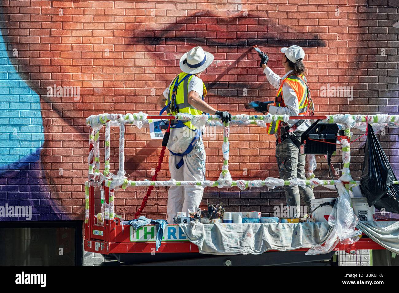 Deux artistes peignent une murale colorée sur un bâtiment en briques tout en se tenant debout sur un ascenseur hydraulique. Centre-ville de Calgary, Alberta, Canada. Banque D'Images