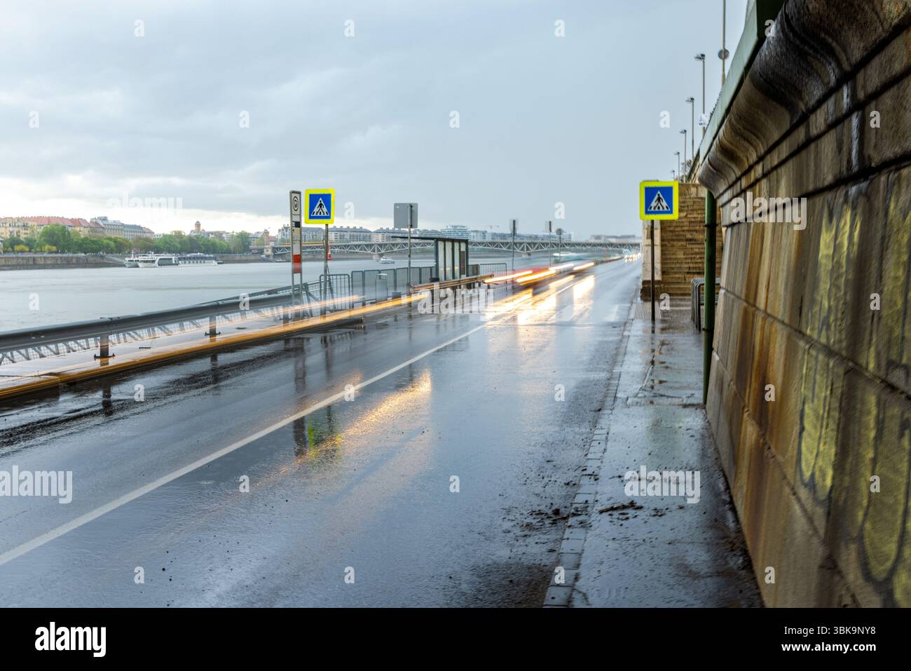 Route urbaine pluvieuse en bord de rivière avec des sentiers de lumière floue des voitures, des panneaux de passage pour piétons et le ciel couvert se reflétant sur la chaussée mouillée Banque D'Images