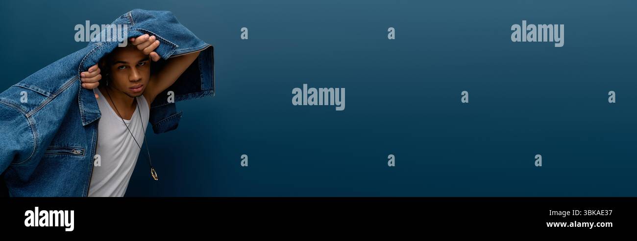 Élégant jeune homme afro-américain pose en toute confiance dans une veste en denim sur un fond bleu Uni, bannière Banque D'Images