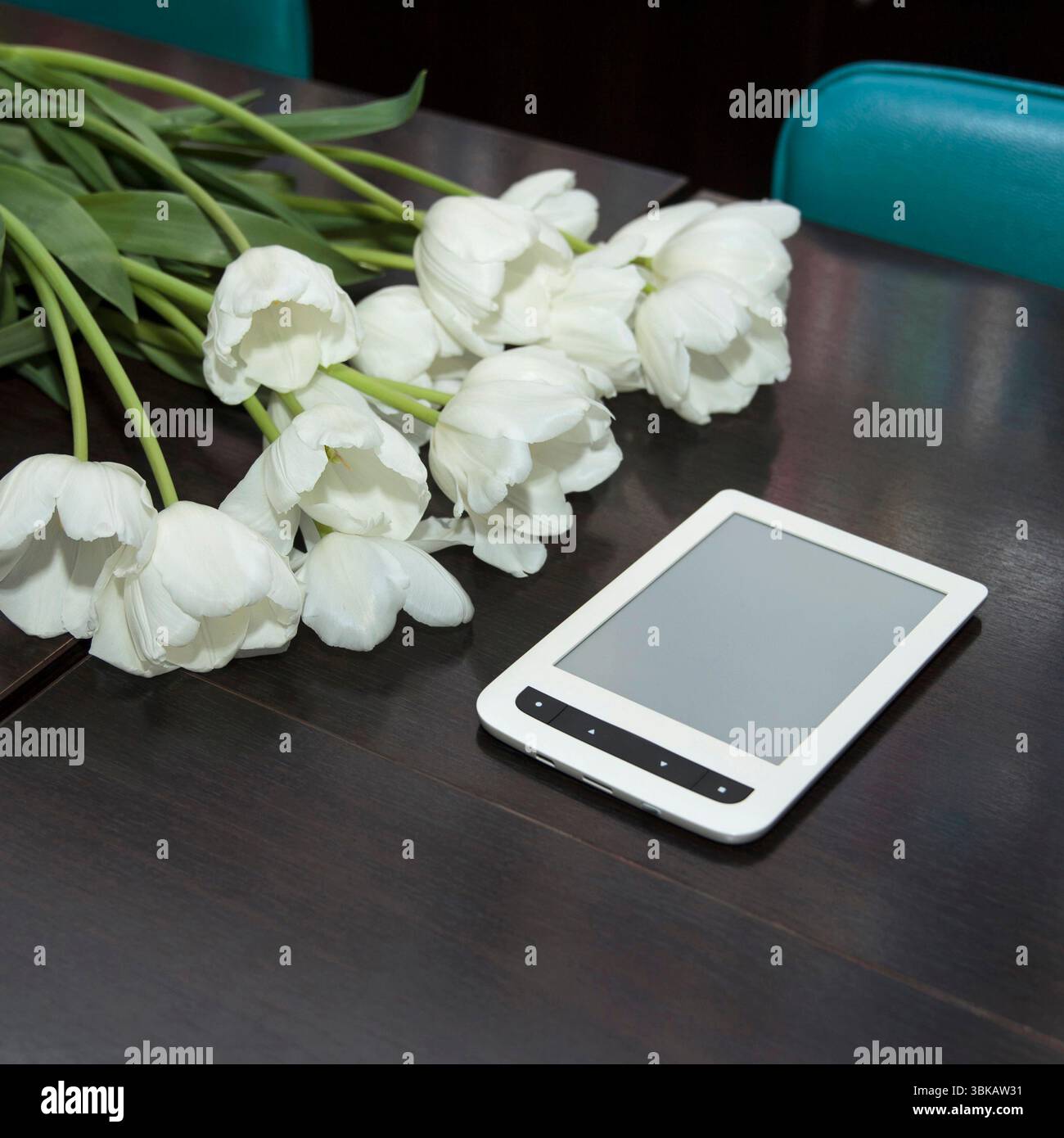 Un bouquet de tulipes blanches et un lecteur électronique blanc sur une table en bois sombre, entouré de chaises, dans le public Banque D'Images