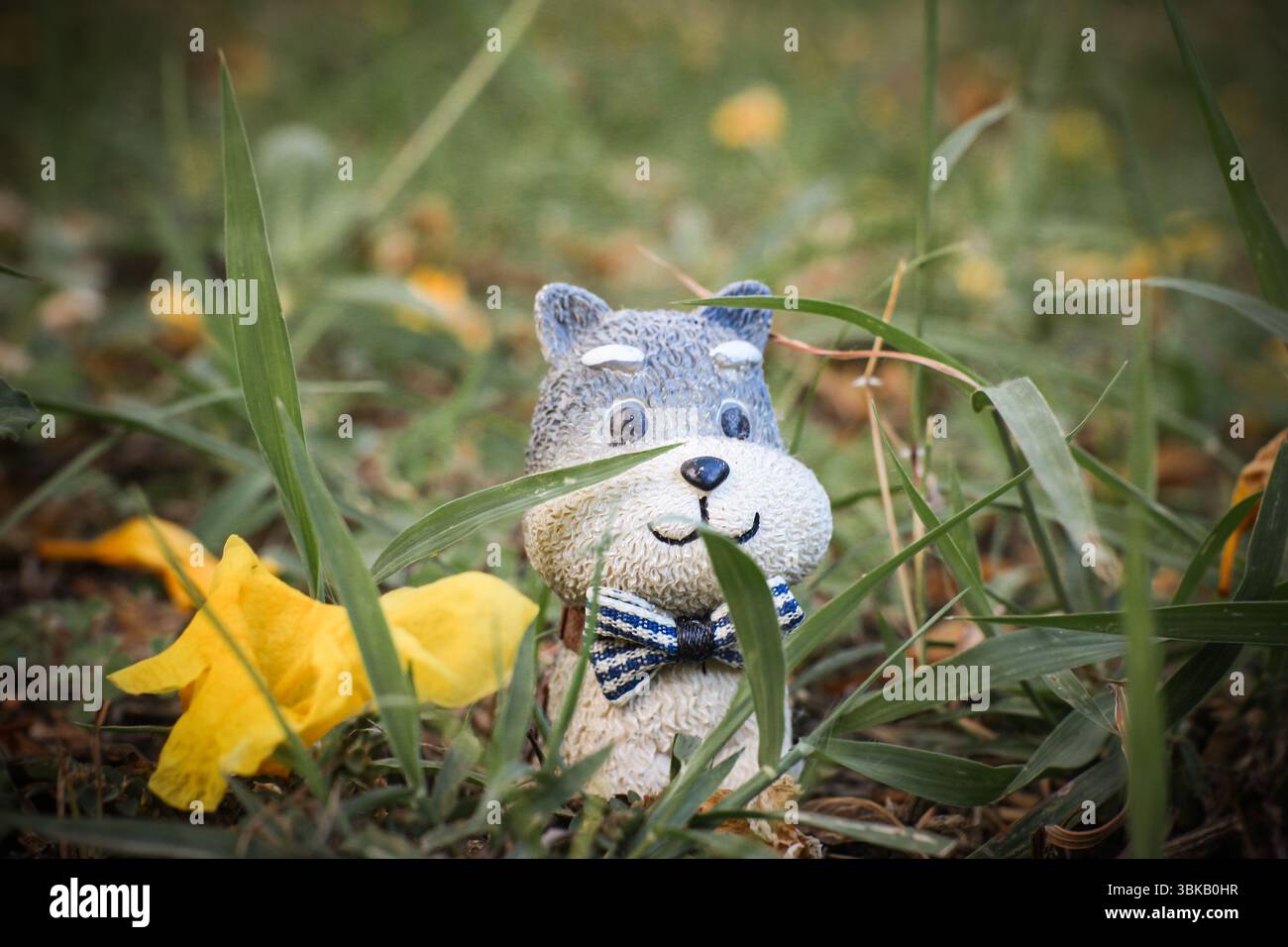 Figurine miniature de chien en argile avec un noeud papillon parmi les fleurs tombées et l'herbe, capturée dans un fond vert macro. Thème nature et bonheur. Banque D'Images