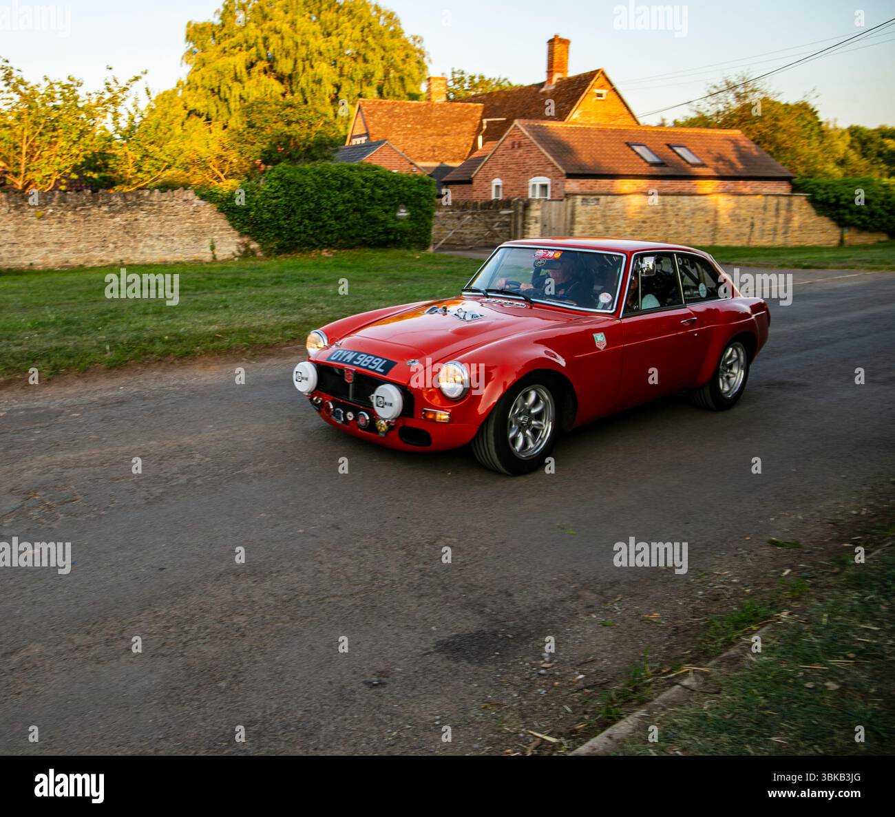Une voiture rouge classique MGB GT roule le long d'une route rurale devant un mur de pierre et des maisons de briques sous la lueur dorée du soleil couchant Poulshot Devizes Banque D'Images