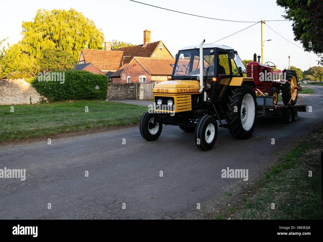 Tracteur Leyland 802 remorquant une machine agricole rouge antique sur une route rurale avec des maisons traditionnelles en briques en arrière-plan Banque D'Images