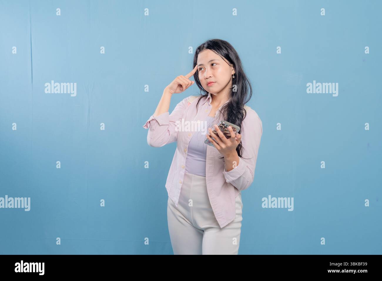 Une femme est debout devant un mur bleu, regardant vers le bas et pensant. Elle porte une chemise blanche et un pantalon blanc Banque D'Images
