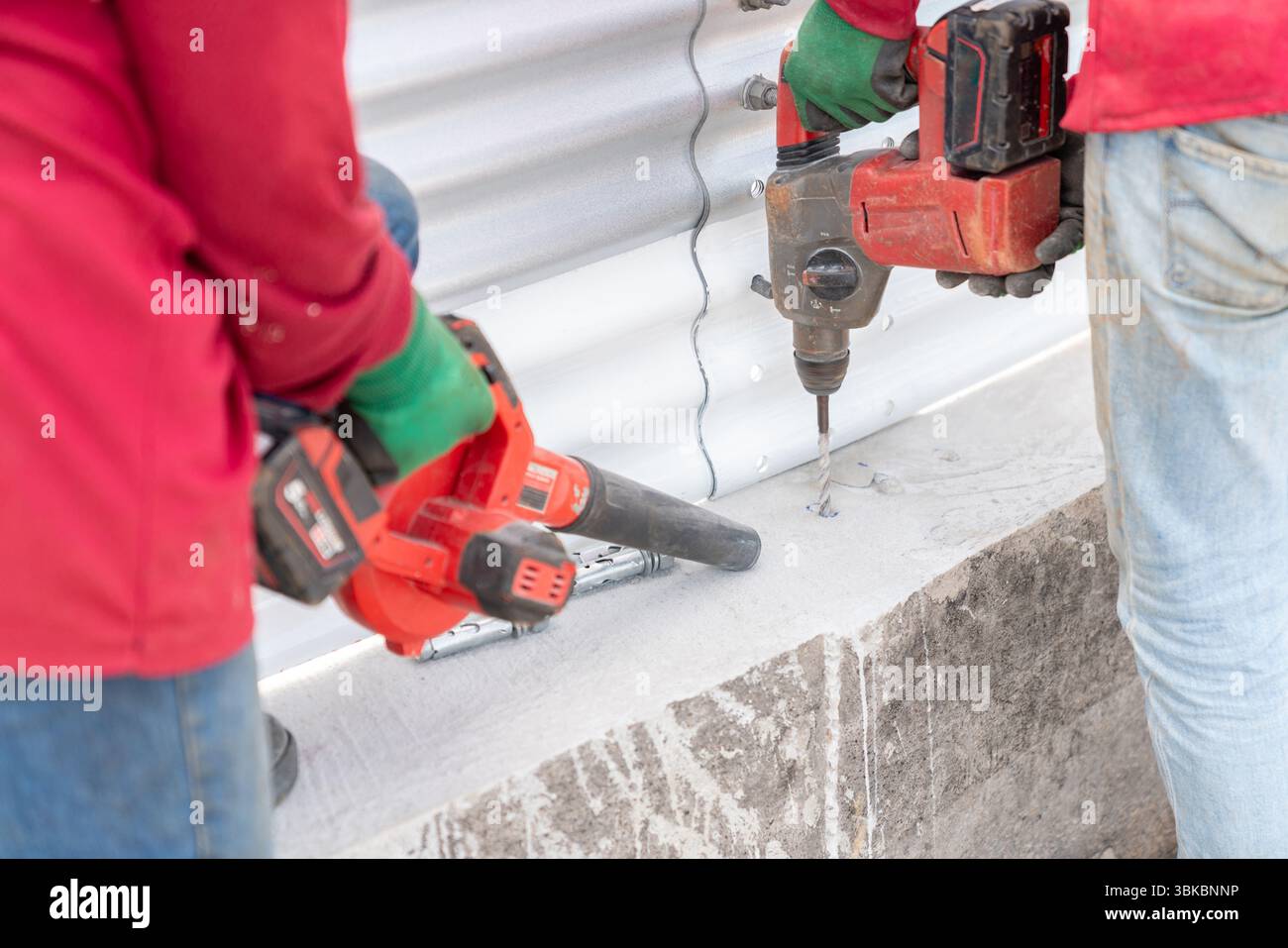 Deux ouvriers forent dans le plancher de béton avec une perceuse sans fil et utilisent un ventilateur sans fil pour enlever la poussière et les débris de la zone de travail. Banque D'Images