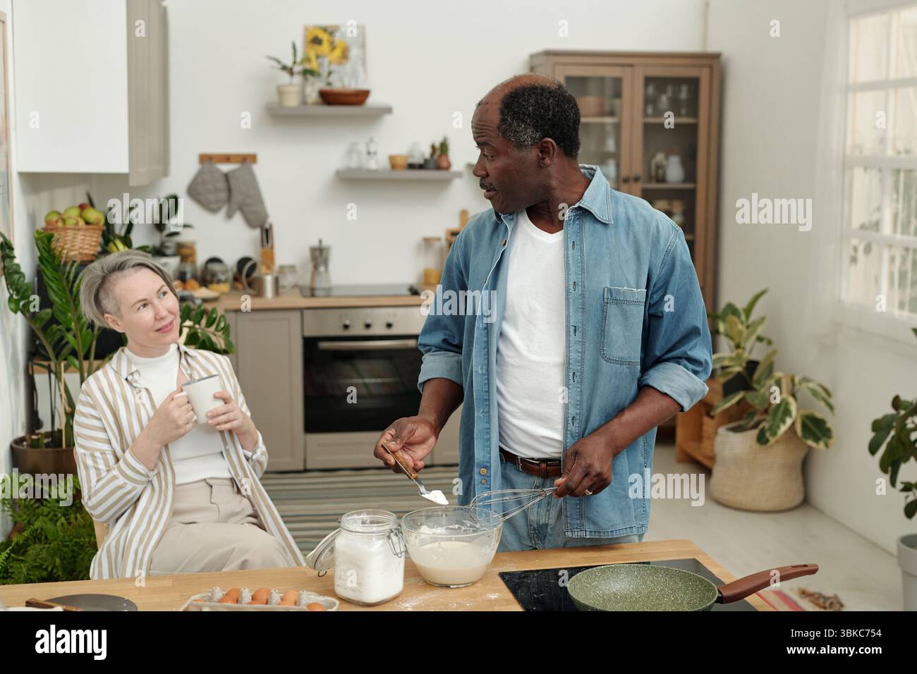 Couple multiethnique préparant le petit déjeuner dans une cuisine moderne, entouré de plantes et d'ustensiles de cuisine, partageant un moment de conversation et de rire Banque D'Images
