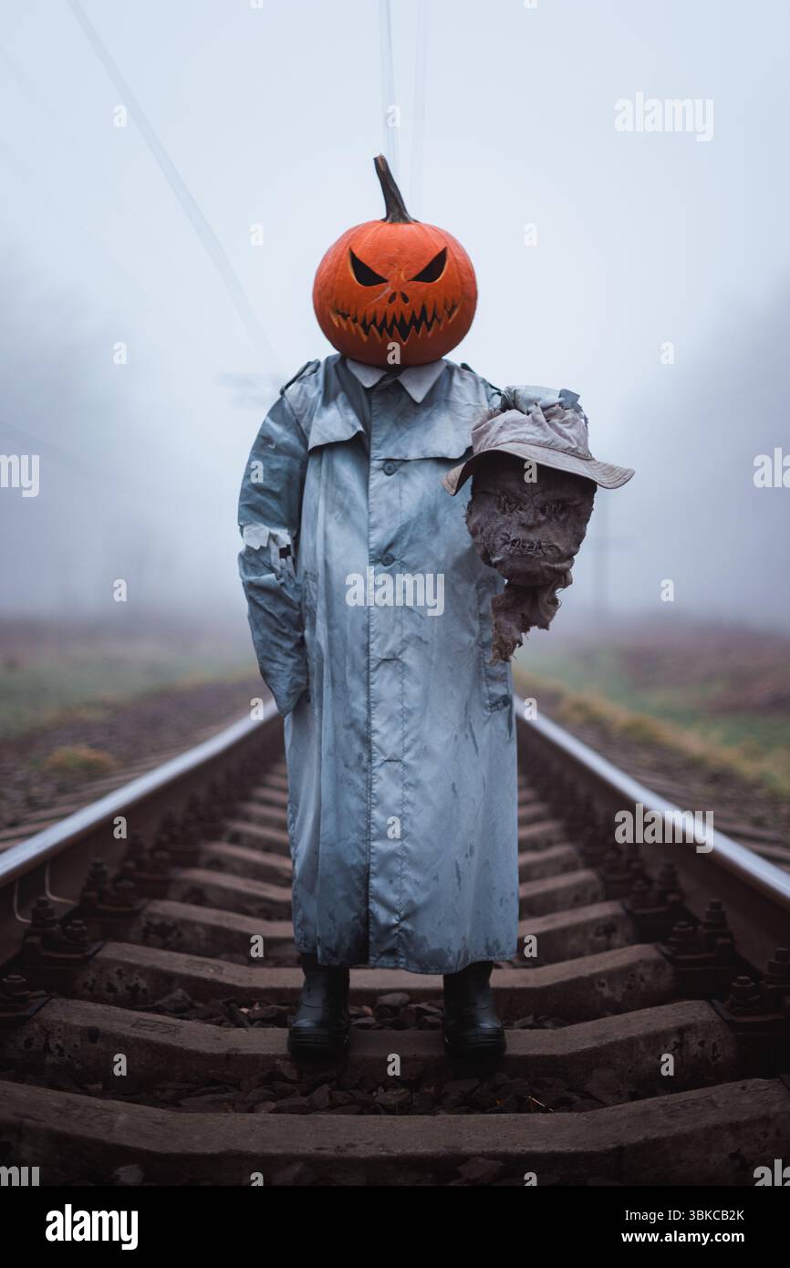 Figure effrayante dans le masque de citrouille sculpté avec la tête coupée d'épouvantail dans la main se tient sur le chemin de fer brumeux dans la campagne d'automne. Concept d'Halloween Banque D'Images