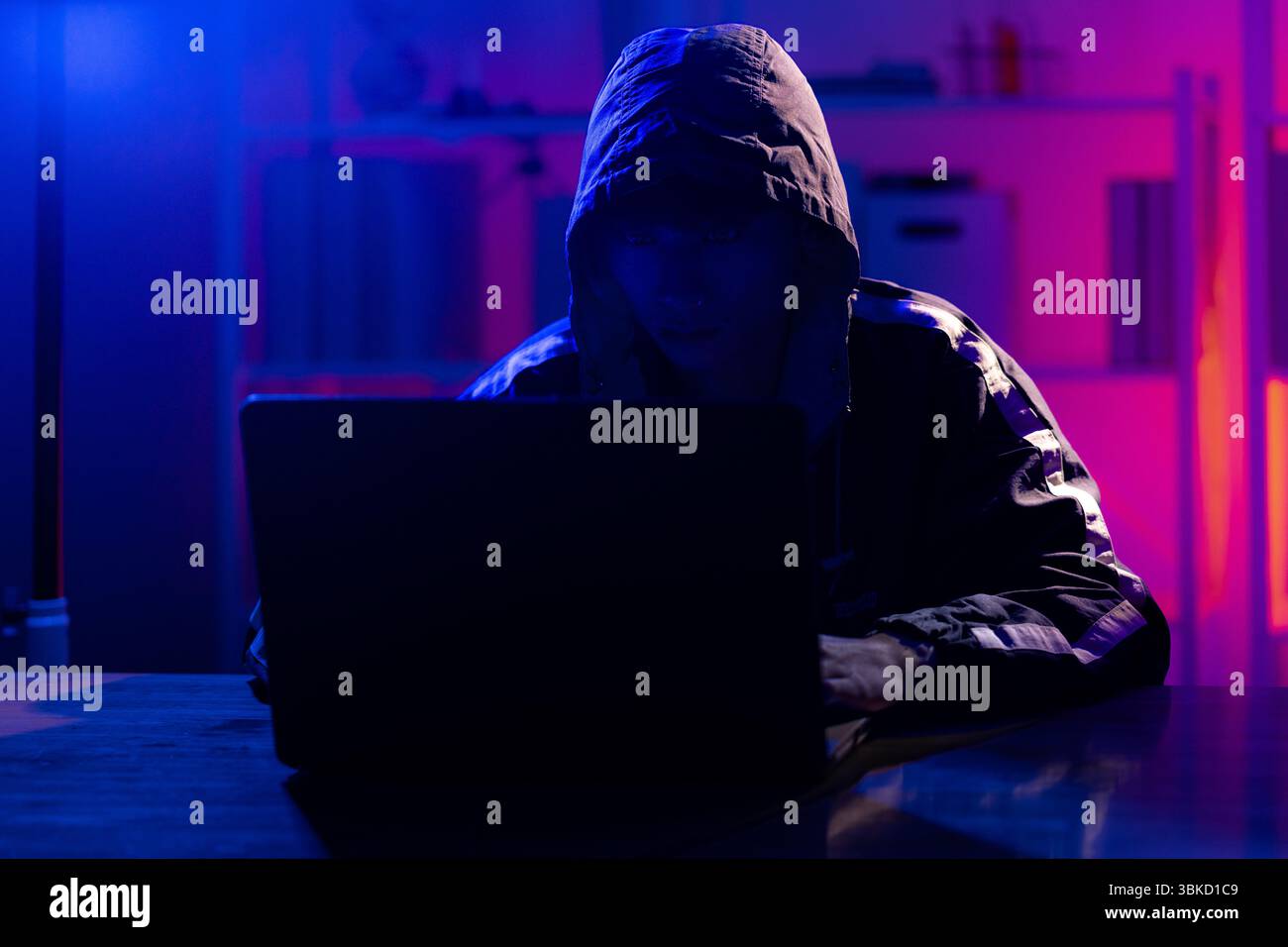 Un jeune homme portant un sweat à capuche noir utilisant intentionnellement un ordinateur portable dans une pièce éclairée au néon, évoquant les thèmes de la cybersécurité, du piratage, du codage et de la confidentialité numérique dans un t Banque D'Images