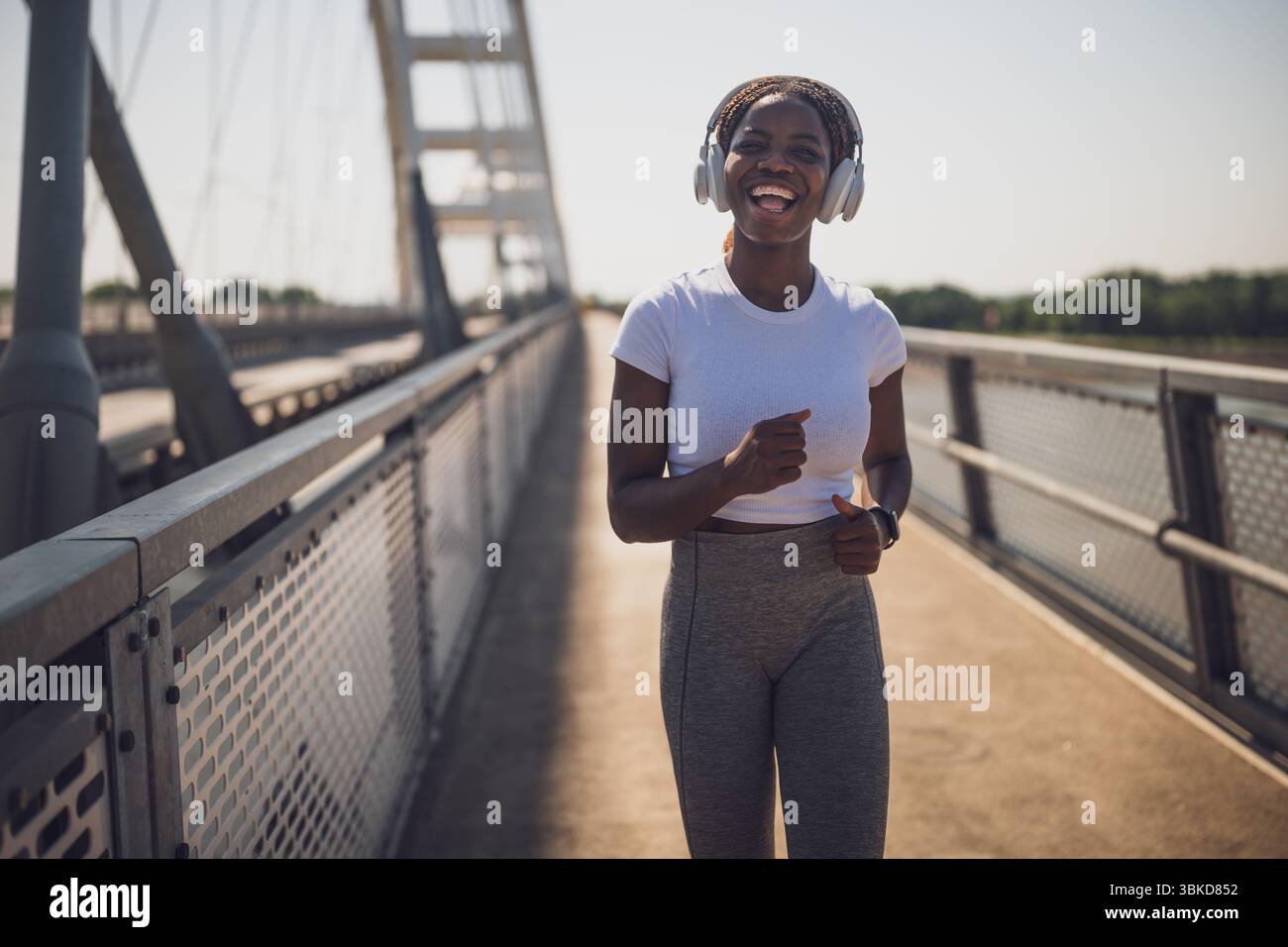 Femme afro-américaine sportive heureuse et en forme dans le jogging sportswear et écouter de la musique à travers des écouteurs sur le pont de la ville urbaine. Banque D'Images