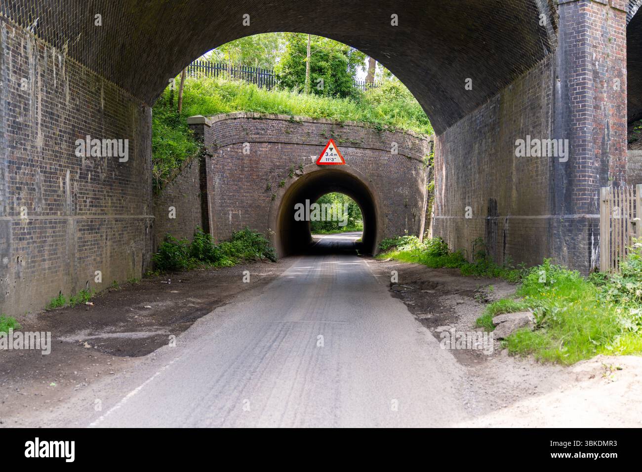Route asphaltée serpentant sous un pont de briques, menant à un tunnel sombre marqué par un panneau de limite de hauteur, créant un passage intrigant Banque D'Images