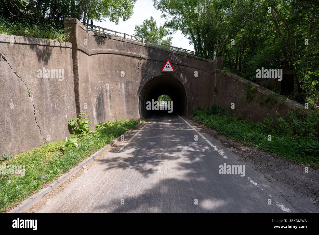 Route de campagne tranquille passant sous un pont bas avec un panneau de limite de hauteur, entourée d'une végétation luxuriante Banque D'Images