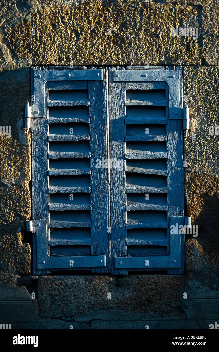 La lumière du soleil projette des ombres sur une paire de volets bleus altérés attachés à un vieux mur de pierre. Auvergne, France Banque D'Images