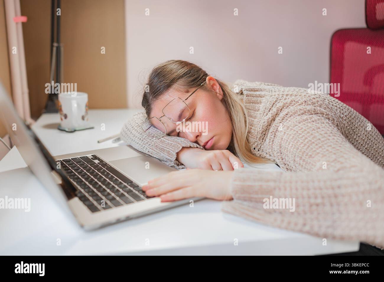Femme dormant au bureau à la maison avec la tête sur les bras à côté de l'ordinateur portable. Position de repos profonde avec les lunettes inclinées et les yeux fermés, chaise rouge, tasse et stylo sur W propre Banque D'Images
