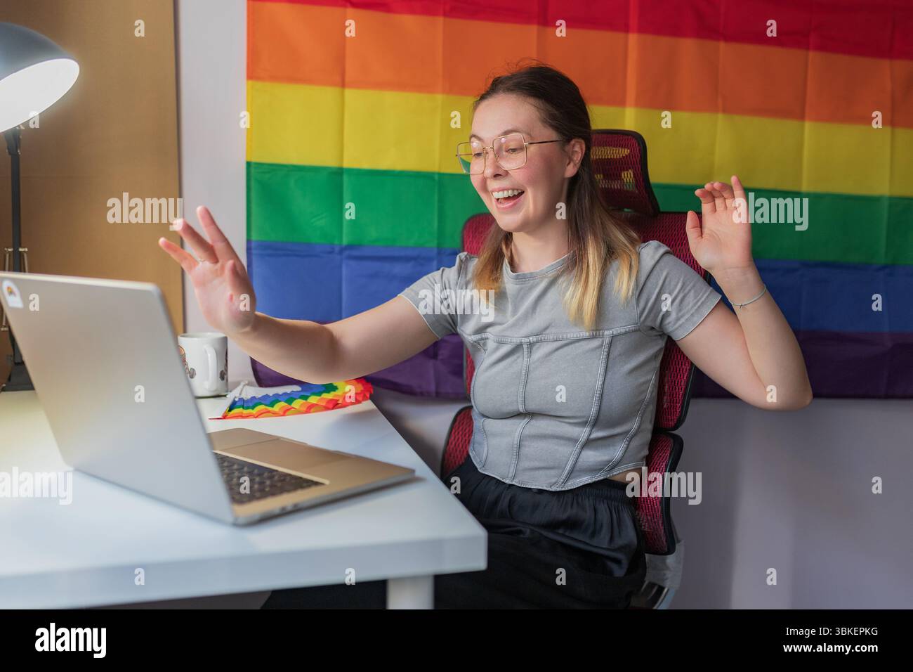 Heureuse femme saluant pendant la réunion vidéo, drapeau arc-en-ciel toile de fond visible / souriante jeune femme levant la main devant l'ordinateur portable, assise au bureau avec RA Banque D'Images
