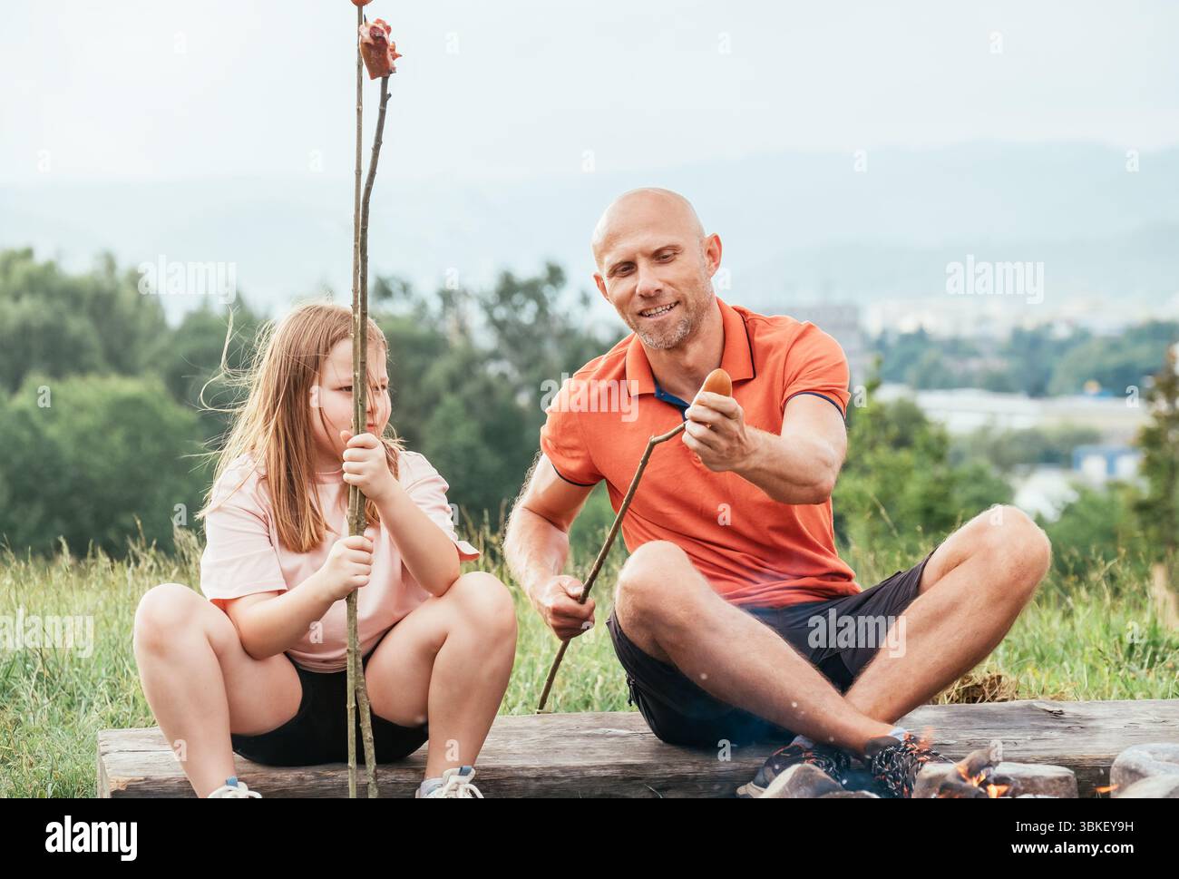 Scène extérieure de famille mignonne du père montrant sa fille comment mettre la saucisse sur le bâton de barbecue alors qu'ils sont assis à côté d'un feu de camp pendant la randonnée dans la nature e Banque D'Images