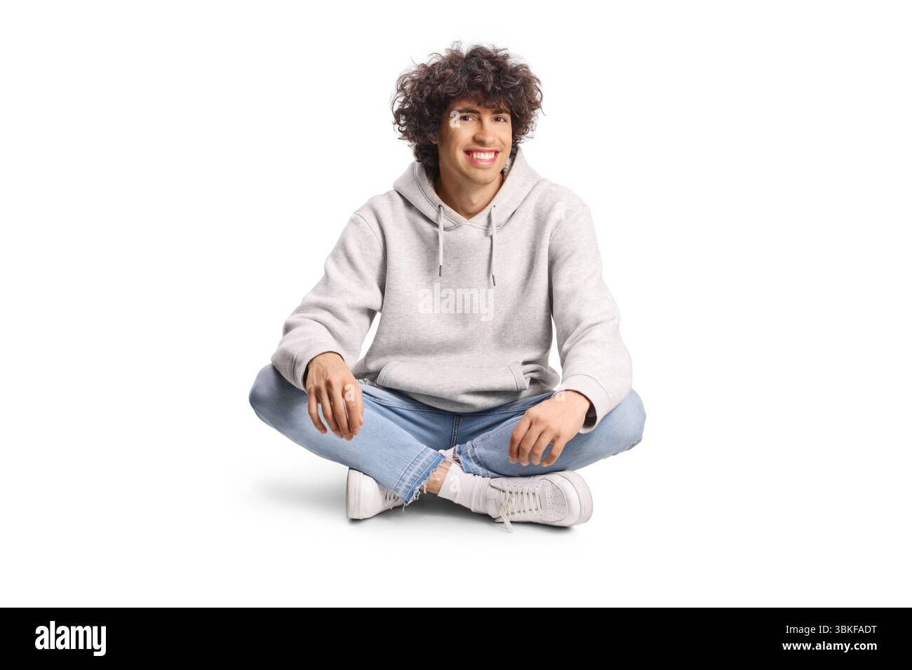Jeune homme aux cheveux bouclés assis avec les jambes croisées isolé sur fond blanc Banque D'Images