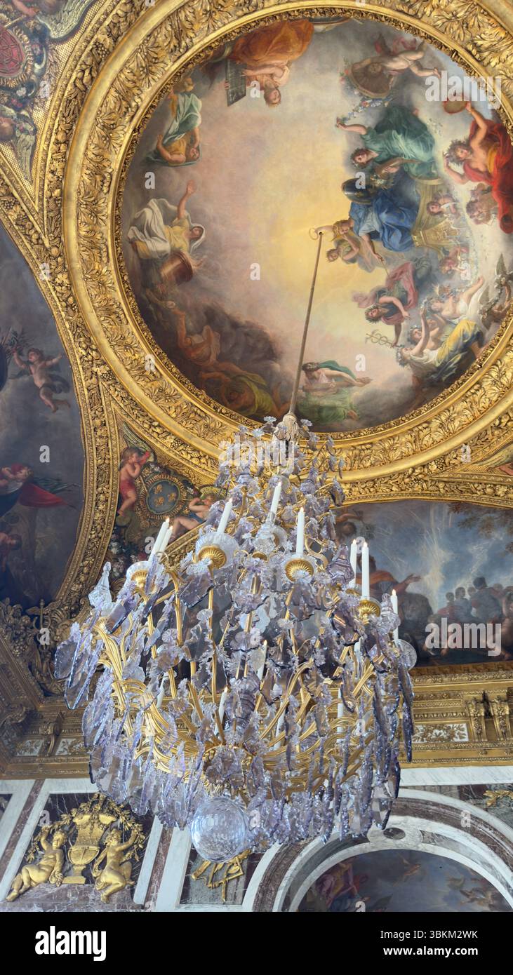 Lustre orné dans une grande pièce. Un plafond peint représente des figures. Couleurs : or, blanc, bleu et rose, avec de riches détails architecturaux. Banque D'Images