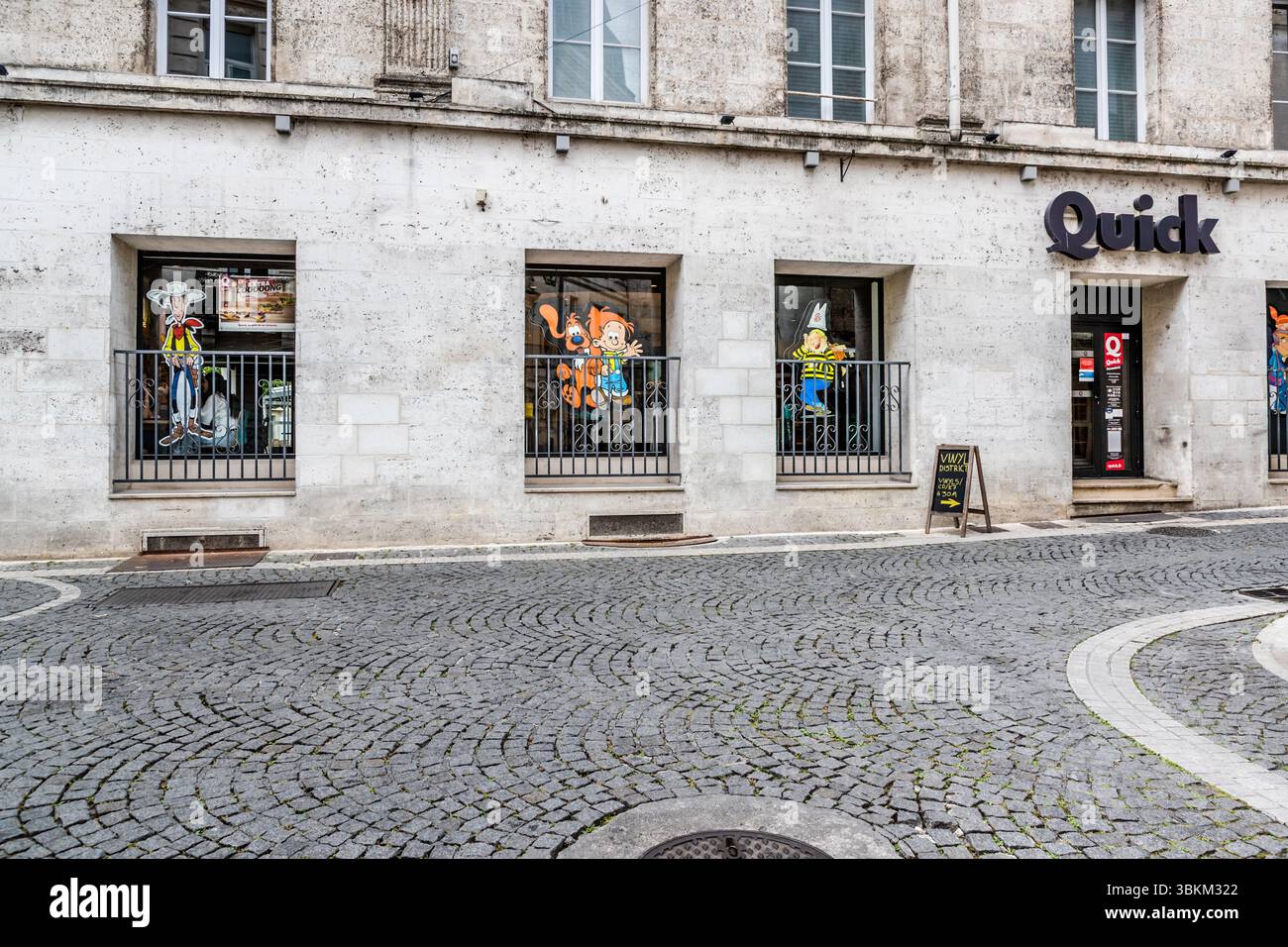 Des personnages de bandes dessinées décorent les fenêtres d'un restaurant Quick dans un bâtiment en pierre pâle, Boulevard Pasteur, Angoulême, Nouvelle-Aquitaine, France. À gauche se tient le cow-boy Lucky Luke de l'artiste belge Morris, au centre Boule et son chien Bill de la série "Boule et Bill" de Jean Roba. La fenêtre de droite montre une figure avec une casquette blanche pointue et une chemise rayée jaune et noire. Un panneau sur le trottoir indique une boutique de vinyle à 30 mètres. Des pavés recouvrent la rue devant la façade. La ville est une Ville créative de littérature de l'UNESCO depuis 2019. Banque D'Images