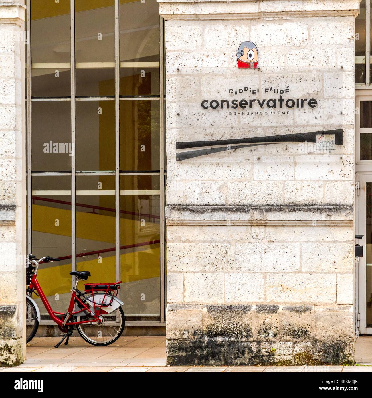 Entrée au Conservatoire Gabriel Fauré (Grand Angoulême). Un vélo rouge se trouve devant le bâtiment historique en calcaire ; une petite figure d'art de rue est apposée sur la façade. Le conservatoire municipal porte le nom du compositeur français Gabriel Fauré (1845-1924), qui a laissé une marque durable dans la région. Conservatoire Gabriel Fauré à Angoulême. Un vélo rouge se tient devant le bâtiment, montrant un escalier jaune à travers une fenêtre. Une œuvre d'art de rue peut être vue sur la façade. Place Henri Dunant, Angoulême, Nouvelle-Aquitaine, France Banque D'Images