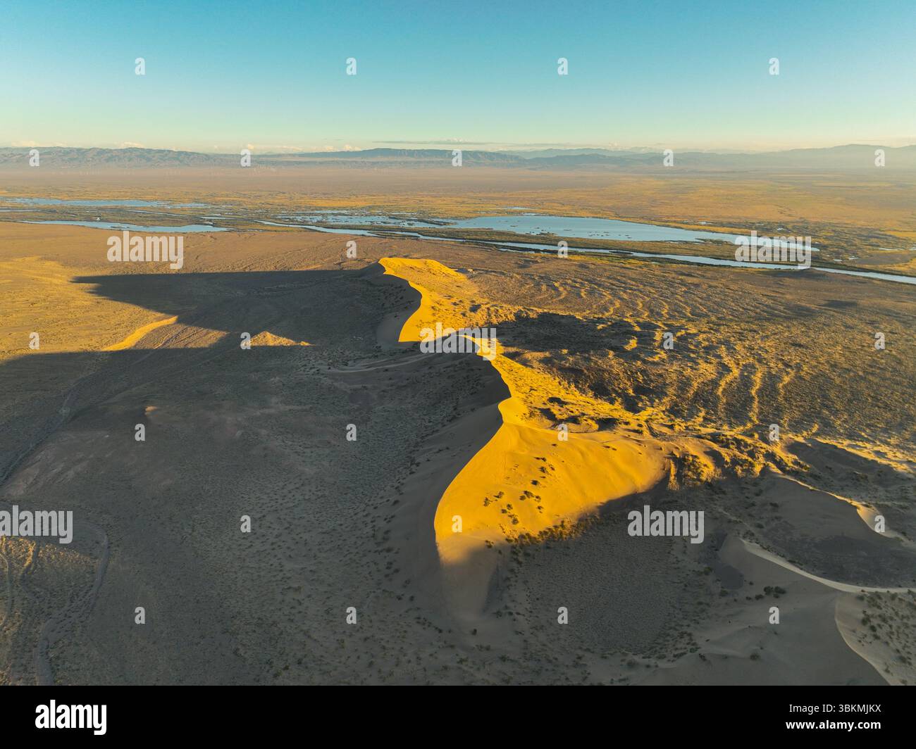 Une vue aérienne capture la forme dramatique de la dune de sable Singing Barkhan projetant une longue ombre à travers le paysage désertique au coucher du soleil, avec w lointain Banque D'Images