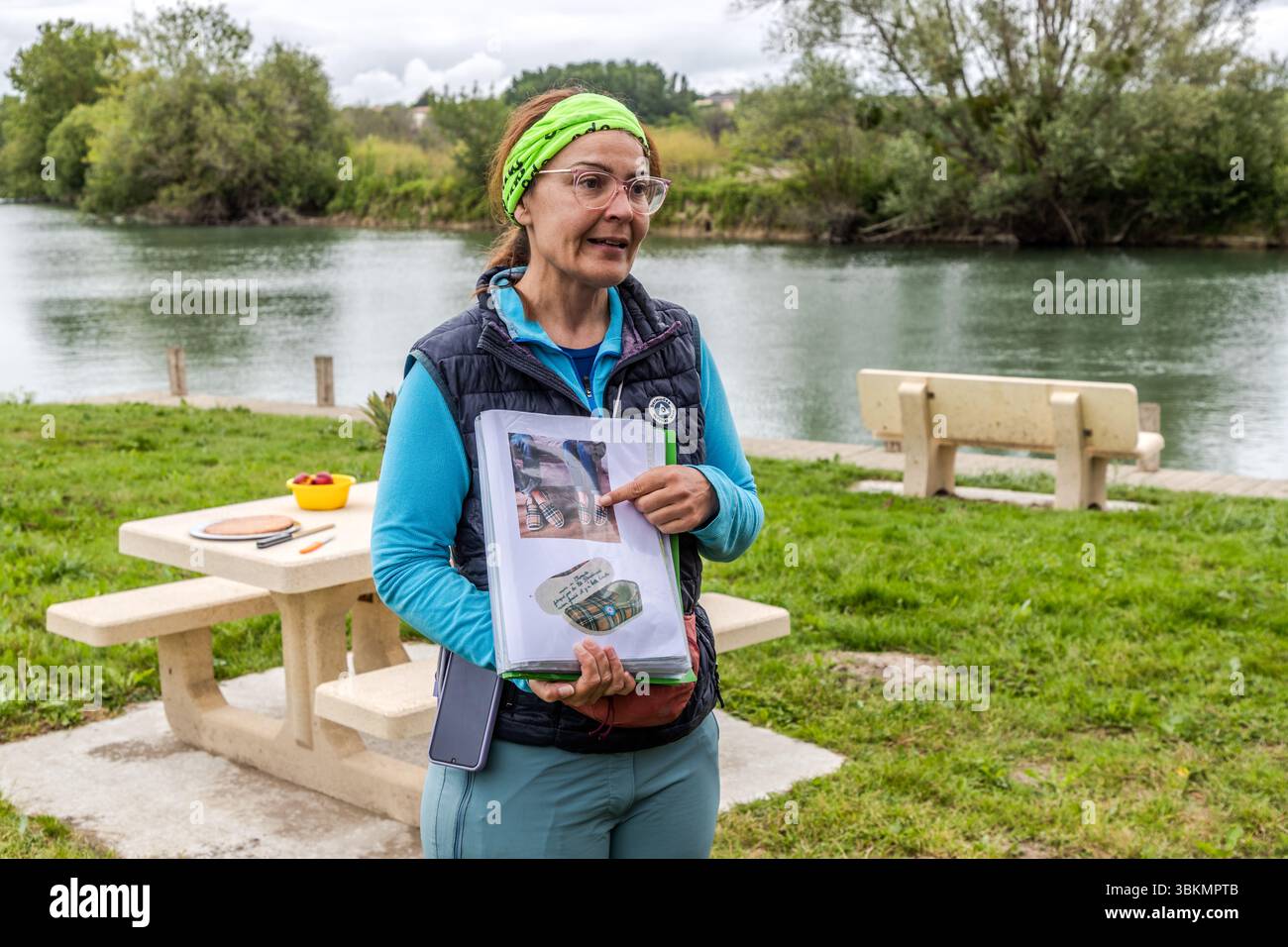 Le guide touristique Katell Nicoux de Die Landpartie informe les participants de la visite à vélo le long de la Charente sur les spécialités régionales. Elle désigne une photo de « Charentaises », des pantoufles emblématiques avec un motif en tissu à carreaux, qui sont fabriquées dans la Charente depuis le XIXe siècle. Katell Nicoux se tient sur la rive de la rivière à Le Ponton, Rouffiac, Nouvelle-Aquitaine, France, à côté d'une table de pique-nique en béton. Le groupe est en visite à vélo guidée sur la route cyclable longue distance La Flow Vélo et utilise la pause pique-nique pour des informations. Banque D'Images
