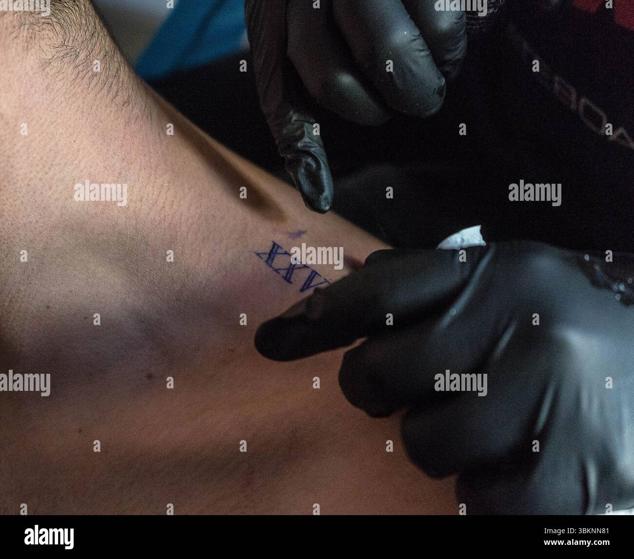 Un tatoueur dans des gants noirs encrage soigneusement un dessin sur le poignet d'une personne. Le tatouage semble être en cours avec l'ajout de chiffres romains Banque D'Images