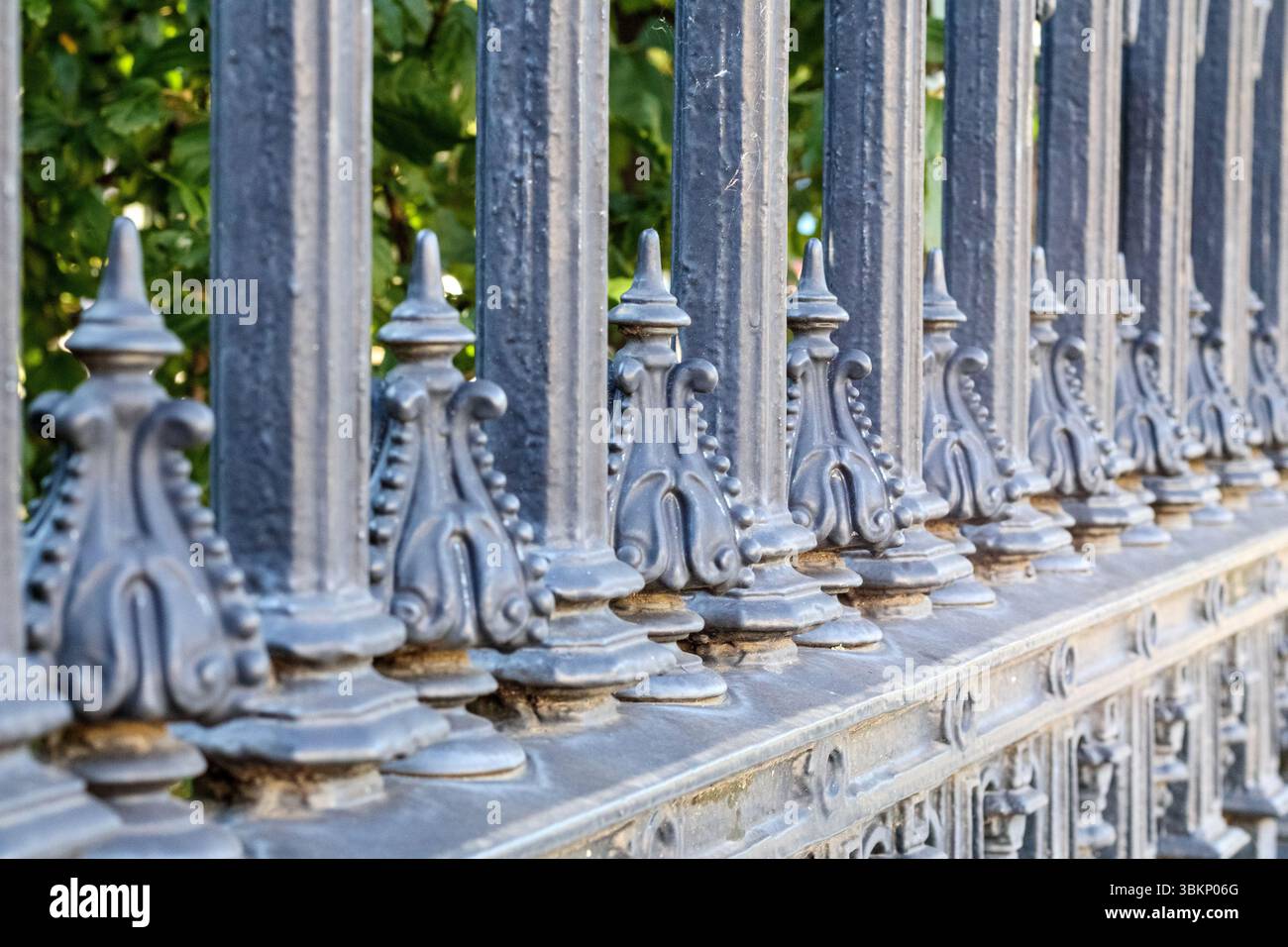 Clôture décorative en métal foncé avec des surfaces pointues et des éléments ornementés, avec des arbres verts et des buissons dans un fond flou. Banque D'Images