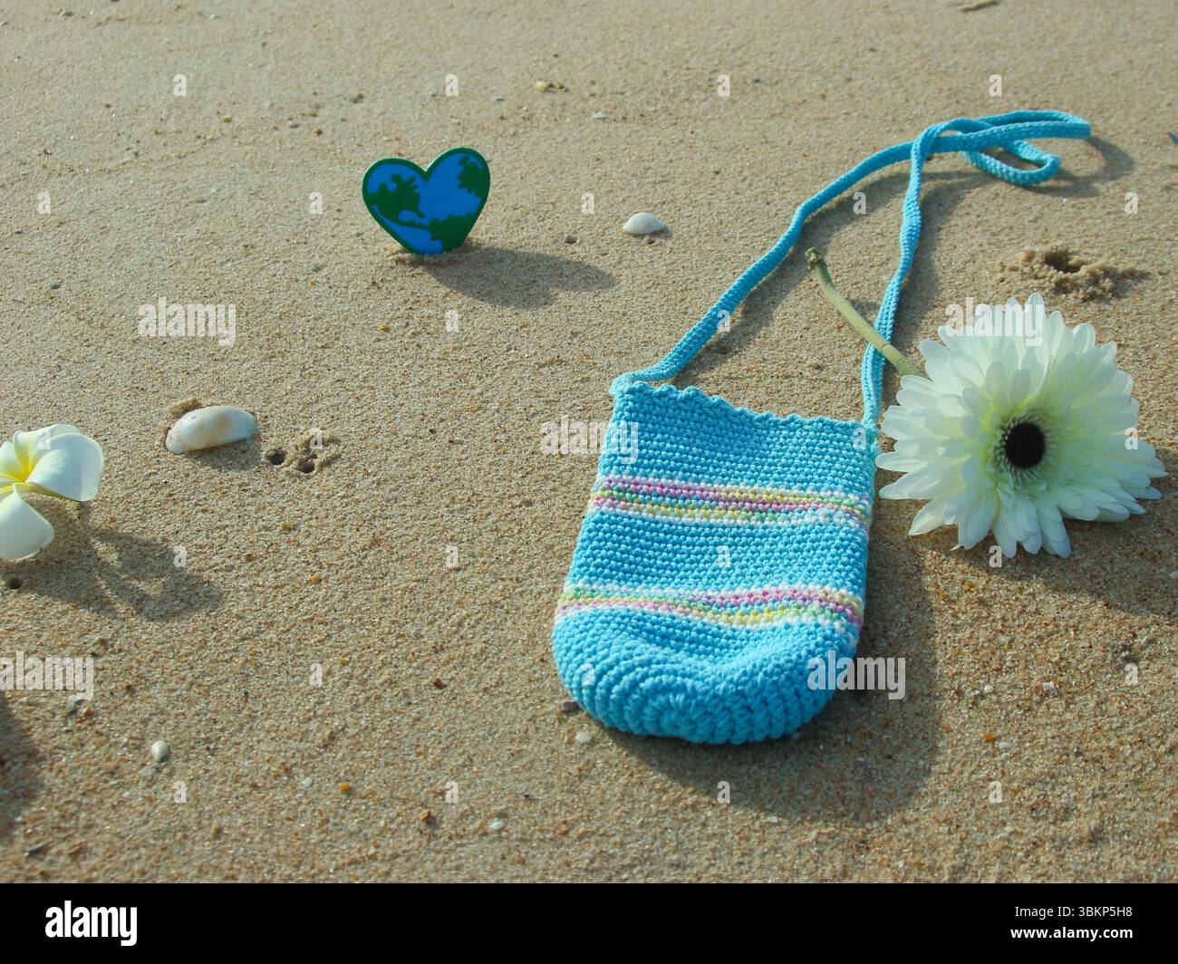Scène de plage écologique avec gerbera blanc, sac au crochet pastel, coquillages, pétales de fleurs et coeur de globe sur sable ensoleillé. Banque D'Images