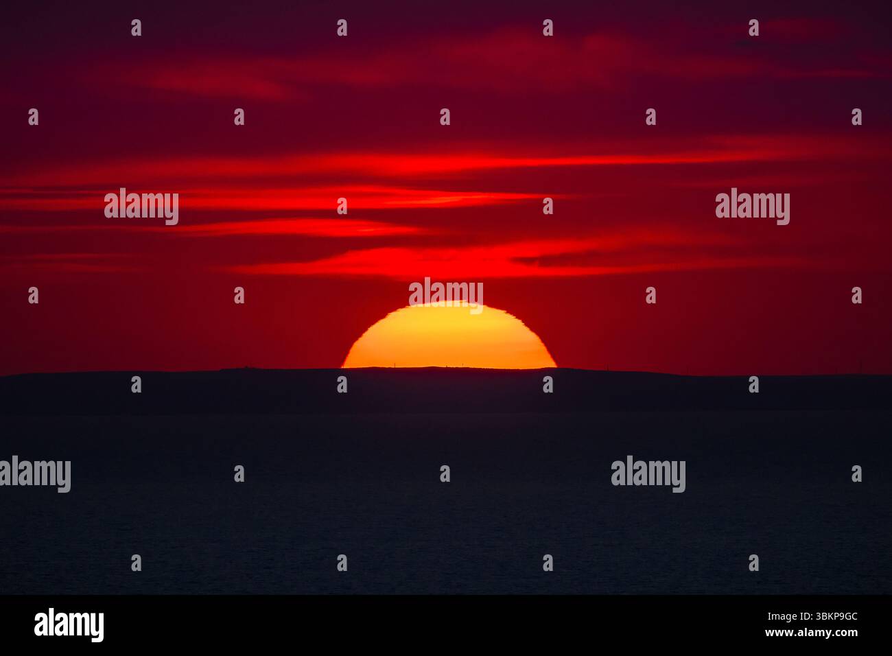 Coucher de soleil rouge vibrant sur l'horizon de la mer avec le soleil à moitié caché derrière une silhouette sombre de collines lointaines. Banque D'Images