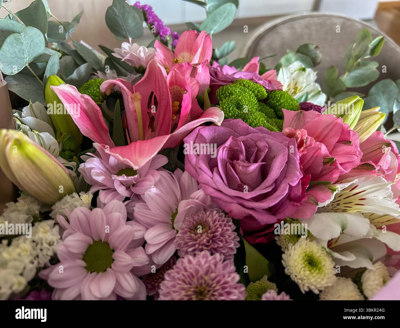 Un gros plan vibrant d'un bouquet printanier avec des lis roses, des roses violettes, des marguerites, des chrysanthèmes verts et des feuilles d'eucalyptus, parfait pour célébrer - Image de stock capturée avec un smartphone