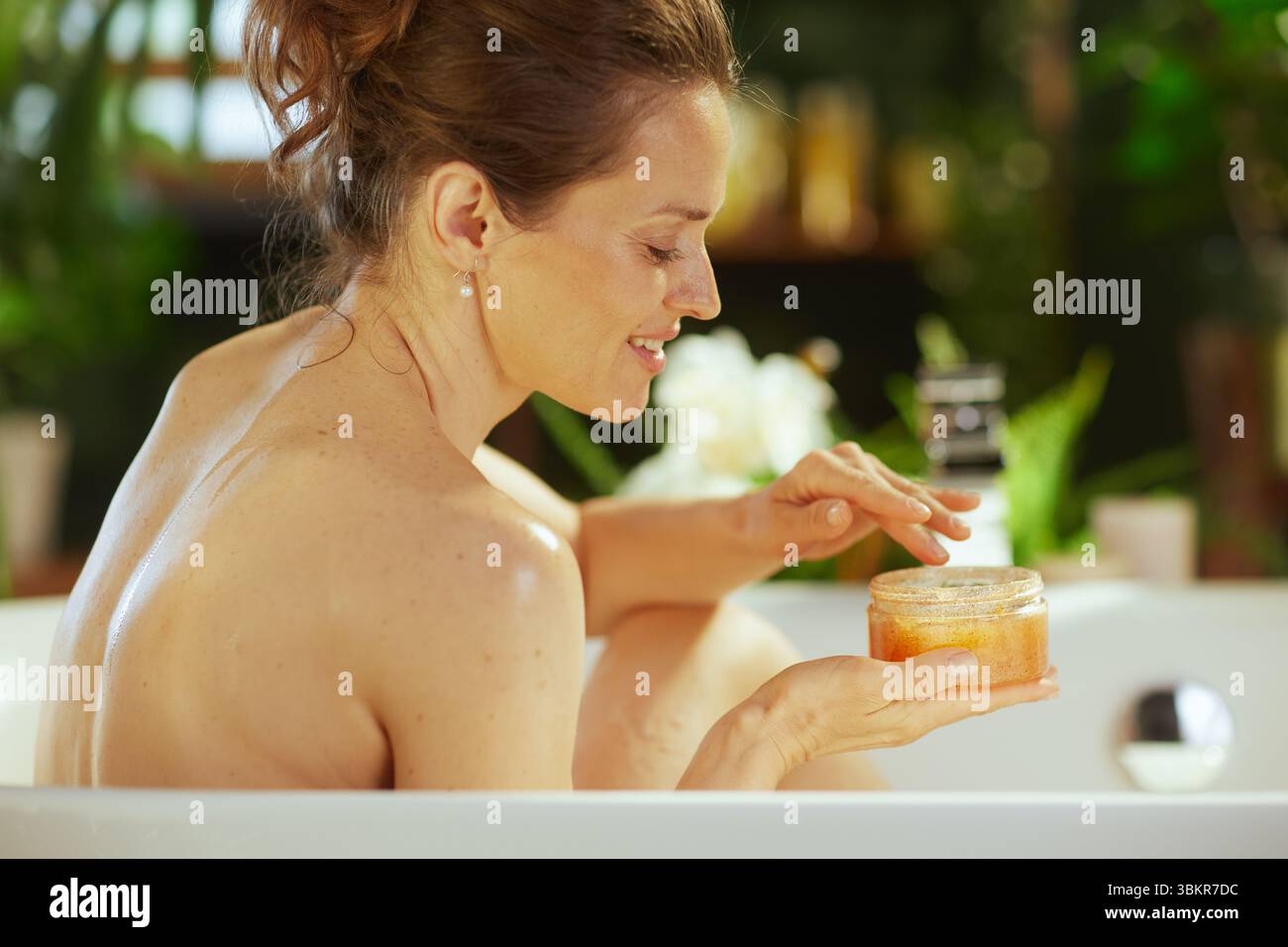 Une femme adulte avec un doux sourire se livre à un rituel d'auto-soin, trempant les doigts dans un gommage corporel tout en se relaxant dans une baignoire blanche immaculée. Tranquille Banque D'Images