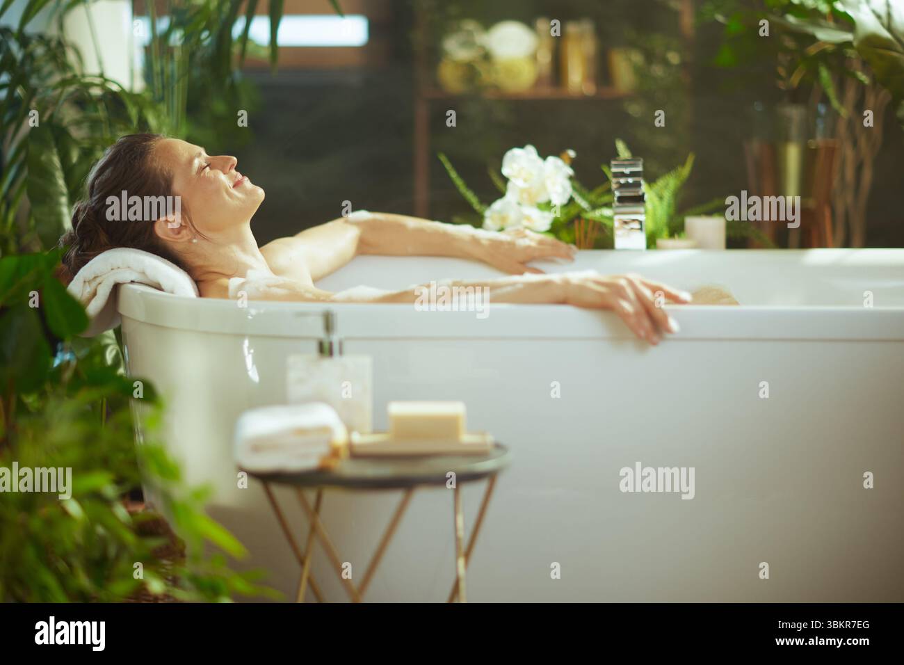 Une femme adulte aux cheveux foncés se détend dans un bain blanc moussant, les yeux fermés avec un sourire serein. Entouré de plantes vertes luxuriantes et d'un éclairage doux, embodi Banque D'Images