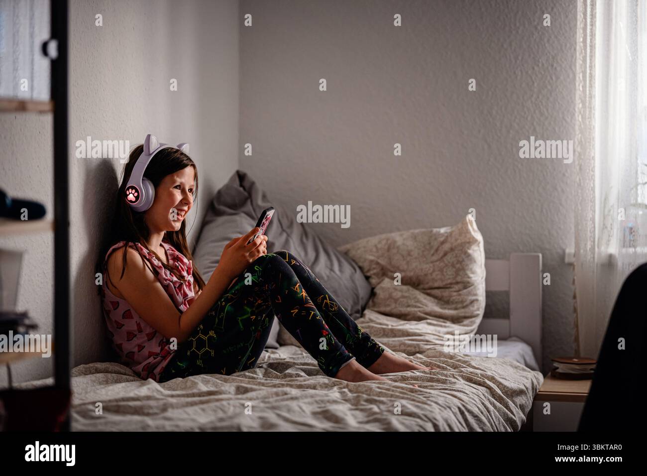 Heureuse fille portant des écouteurs d'oreille de chat lumineux rit tout en utilisant son smartphone, assise confortablement sur un lit confortable près d'une fenêtre. Banque D'Images