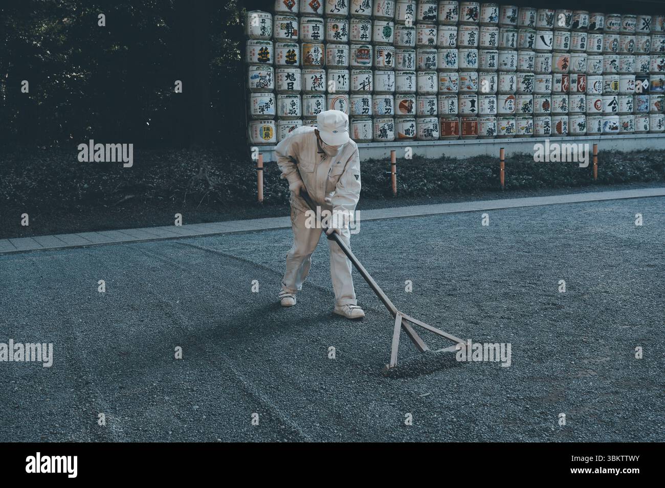 Le gardien du sanctuaire ratisse méthodiquement le gravier devant une exposition vibrante de fûts de saké traditionnels à Meiji Jingu Banque D'Images