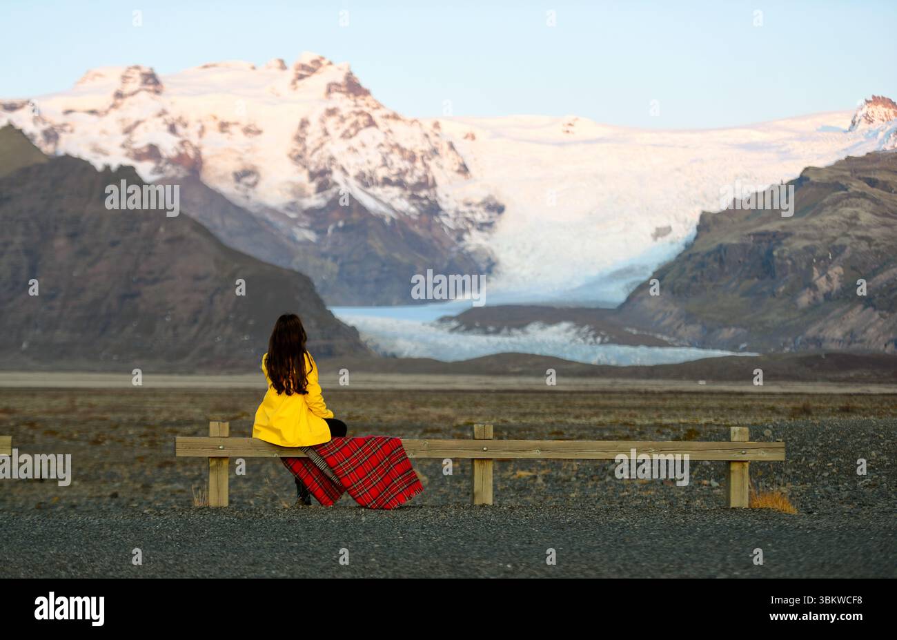 Une femme dans une veste jaune assise sur un banc en bois, regardant le paysage de montagne enneigé. Banque D'Images