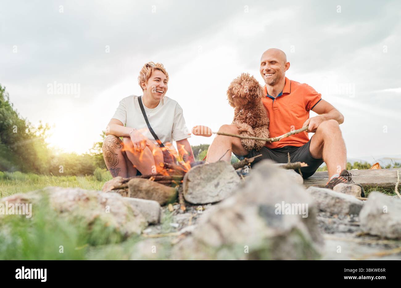 Scène extérieure mignonne de pique-nique : couple souriant heureux avec chien Maltipoo grillant des saucisses sur des bâtons de barbecue assis près d'un feu de camp pendant la randonnée Banque D'Images