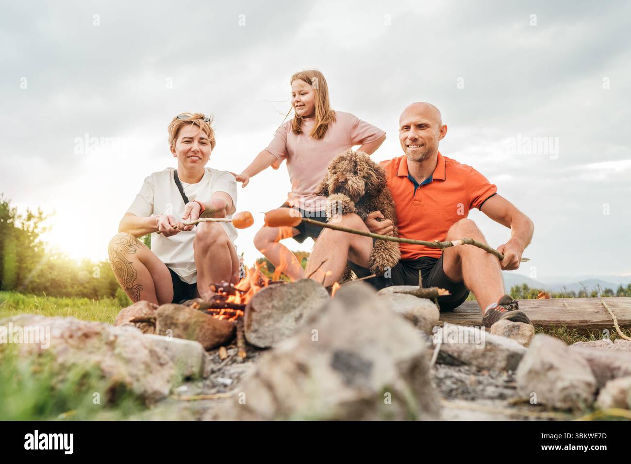 Scène extérieure mignonne de pique-nique : père, fille et maman avec chien Maltipoo griller des saucisses sur des bâtons de barbecue assis près du feu de camp pendant la randonnée R Banque D'Images