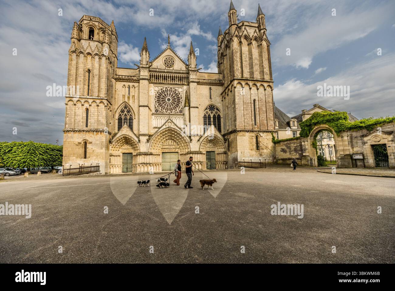 La façade ouest de la cathédrale Saint-Pierre (Cathédrale Saint-Pierre de Poitiers) s'élève sur la Place de la Cathédrale à Poitiers, en Nouvelle-Aquitaine, en France. Deux tours inégales encadrent la façade avec ses trois portails et sa grande rosace. Devant la cathédrale, un homme et une femme promènent leurs chiens en laisse sur la place. À droite, une porte couverte de lierre mène à une cour. La construction a commencé en 1166 à l'initiative d'Eleanor d'Aquitaine et d'Henry II Plantagenêt. À l'époque, ils étaient roi et reine d'Angleterre et ducs de Normandie et d'Aquitaine. Ils étaient d'importants mécènes de Banque D'Images