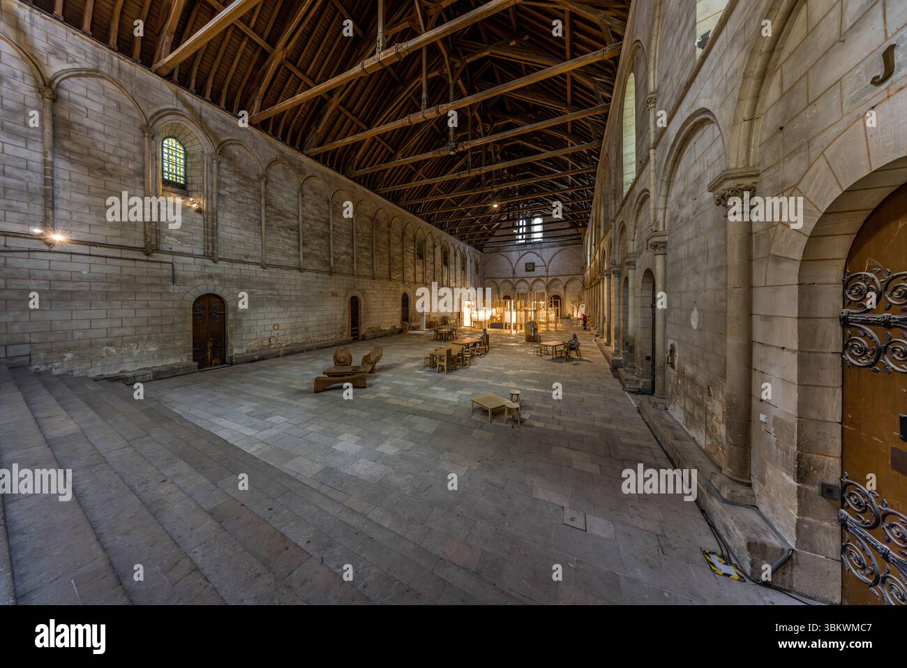 La Salle des Pas Perdus (Hall of Lost Steps) du Palais des ducs d'Aquitaine sur la Rue Gambetta à Poitiers, Nouvelle-Aquitaine, en France, est l'une des plus grandes salles séculières médiévales d'Europe. Un toit en bois ouvert traverse la salle. Des arcades aveugles et de petites fenêtres en arc rond structurent les longs murs. Des tables, des chaises et un banc en bois incurvé se trouvent sur le sol en pierre, et un visiteur est assis à l'une des tables. Des marches en pierre descendent à gauche, et une porte en bois avec des ferronneries se dresse à droite. Trois cheminées monumentales et richement décorées dominent Banque D'Images