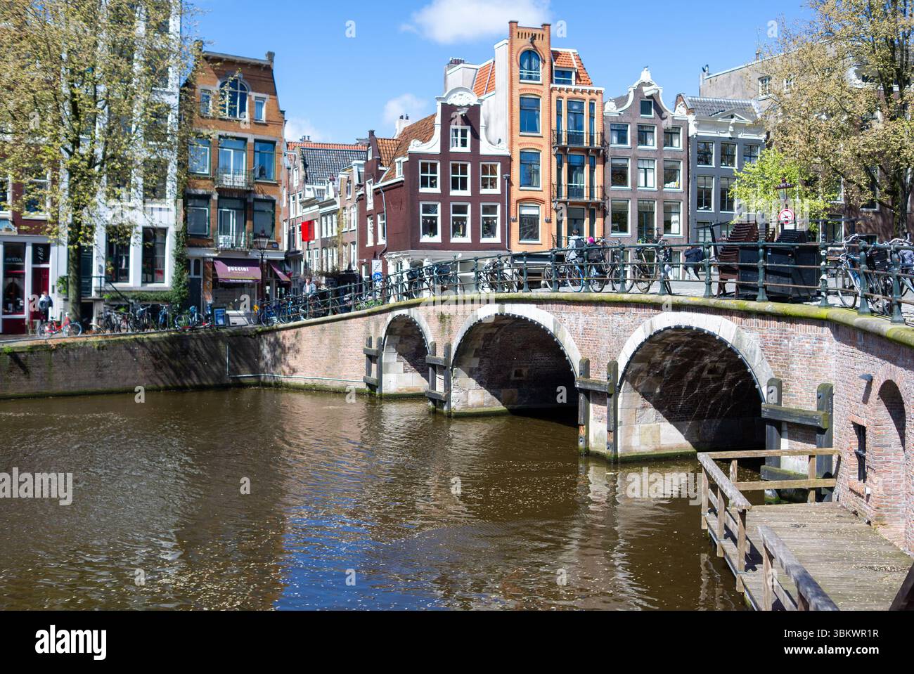 Journée ensoleillée à Amsterdam : les gens se promènent et font du vélo par un pont sur le canal, en profitant de la vue sur les bâtiments hollandais classiques. Banque D'Images