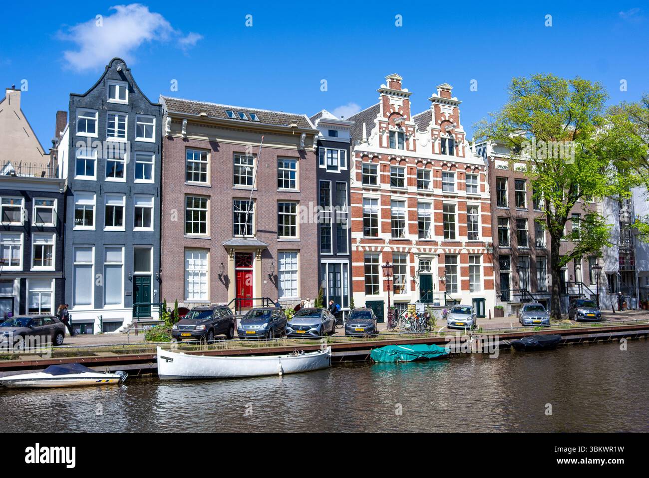 Scène du canal d'Amsterdam : maisons historiques, voitures garées, bateaux et personnes profitant de la journée. Banque D'Images