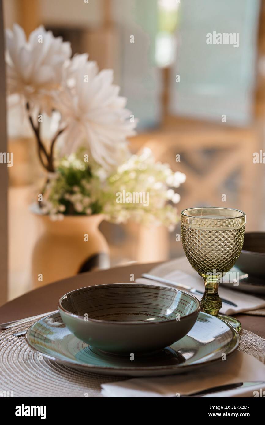 Superbe décor de table avec un verre à vin teinté vert, un bol gris sur des assiettes et un vase orné de fleurs blanches dans un cadre moderne de salle à manger Banque D'Images