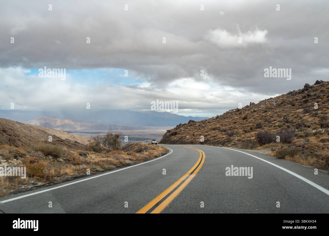 Une route sinueuse traverse un paysage désertique sous un ciel nuageux, avec une voiture seule visible au loin, suggérant un voyage à travers une région isolée et pittoresque. Banque D'Images Une route sinueuse traverse un paysage désertique sous un ciel nuageux, avec une voiture seule visible au loin, suggérant un voyage à travers une région isolée et pittoresque. Banque D'Images