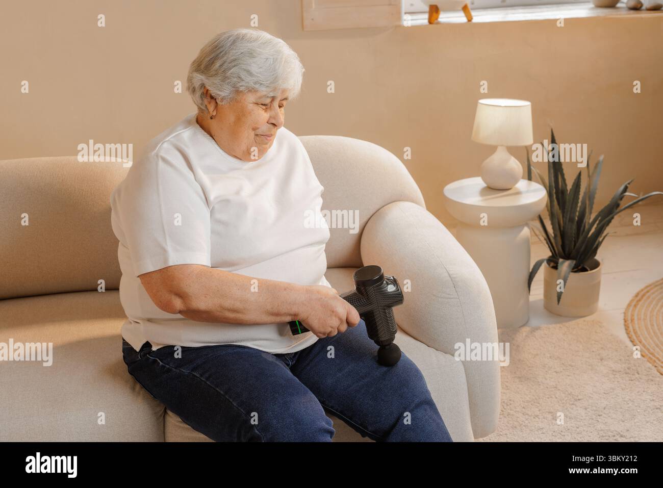 Femme âgée utilisant un pistolet de massage électrique à main sur la jambe pour soulager la douleur thérapie de récupération musculaire et soutien de bien-être à la maison Banque D'Images